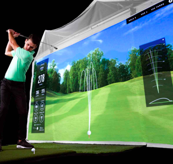 Simulator screen with golf course projected onto screen. Golfer in a swing stance with golf ball on the ground.